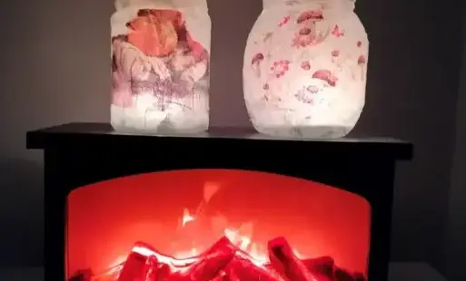 Na zdjęciu są dwa lampiony (jeden ozdobiony grzybkami z serwetki, a drugi dyniami z serwetki) stoją na sztucznym kominku na baterie. W kominku jest płomień. Kominek stoi na białym stoliku. W tle widać białą ścianę.