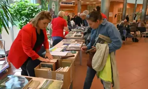 Magdalena Matuszewska rozmawia z czytelnikiem przy stoisku biblioteki. Na stoliku publikacje biblioteki i inne regionalne. Za nimi widać stoliki innych wystawców i zgromadzonych przy nich czytelników