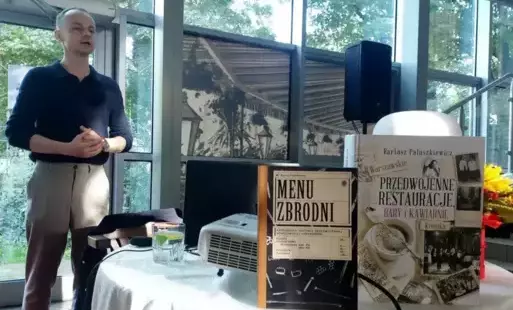 Zdjęcie przedstawia wnętrze jasnego pomieszczenia ze szklanymi ścianami i sufitem, przez które widać drzewa na zewnątrz. Na pierwszym planie widoczny jest stół nakryty białym obrusem. Na stole stoją dwie książki: Jedna z nich nosi tytuł 'Menu zbrodni'. Dr