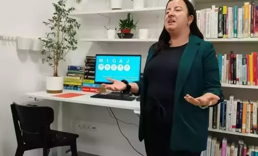 Prowadząca zajęć, Agata Zawadzka pokazuje jak wymigać słowa. Za nią w tle jest monitor z wyświetlonym slajdem prezentacji o miganiu. Monitor stoi na białym biurku, na którym jest też ozdobne drzewko oraz książki. Przy biurku jest czarne krzesło. Nad bi