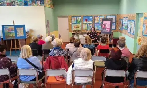 Uczestniczki spotkania siedzą na krzesłach. Przodem do nich siedzi pisarka. Za nią na sztaludze jest plakat informacyjny na sztaludze.