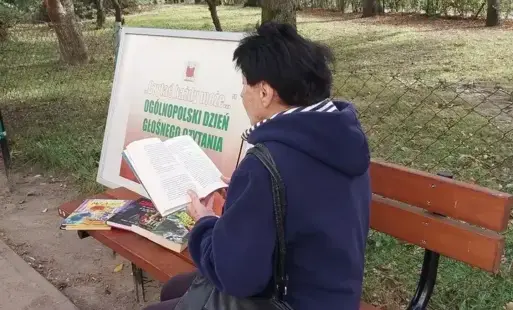 Pani siedzi na ławce i czyta książkę. W tle widoczny park oraz tablica informacyjna.