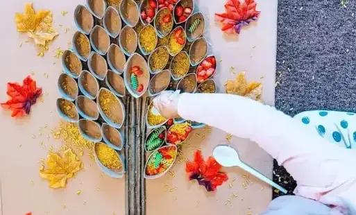 (tytułowe): Dziecko wsypuje do drzewka z rolek czerwoną fasolkę, żółty ryż i zielony makaron, tworząc barwną jesienną kompozycję.