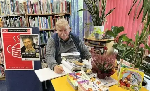 Tytułowe - Mężczyzna siedzący przy stole, na blacie książeczki i kwiat w doniczce