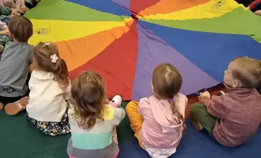 Na dywanie dookoła kolorowej chusty animacyjnej siedzą dzieci.