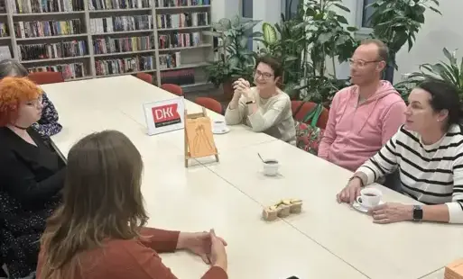na zdjęciu widać uczestników spotkania, którzy siedzą przy dużym, prostokątnym stole. Na stole są egzemplarze omawianej książki, stoją filiżanki z herbatą. Z tyłu znajdują się regały z książkami, a po prawej stronie dwa duże okna. 