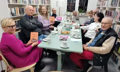 uczestnicy Dyskusyjnego Klubu Książki siedzą przy stole, na którym są książki, przekąski, talerzyki, filiżanki, logo DKK. Po obu stronach widać białe regały z książkami, w tle widać okno.