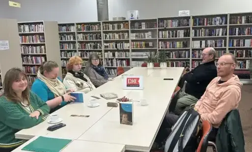 Na zdjęciu widać uczestników spotkania, którzy siedzą przy dużym, prostokątnym stole. Na stole są egzemplarze omawianej książki, stoją filiżanki z herbatą. Z tyłu znajdują się regały z książkami. 