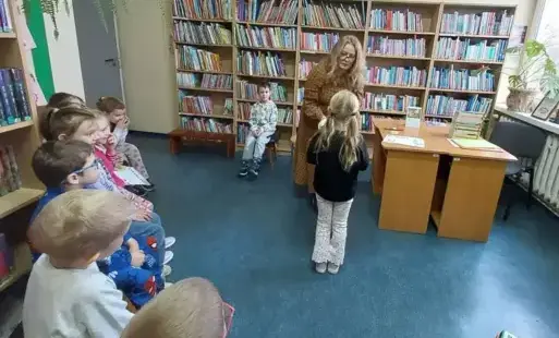 Osoba dorosła, przed nią stojące dziecko, z boku siedzące dzieci. W tle regały z kolorowymi książkami, zielone rośliny, jasno brązowe biurko, na nim książki.