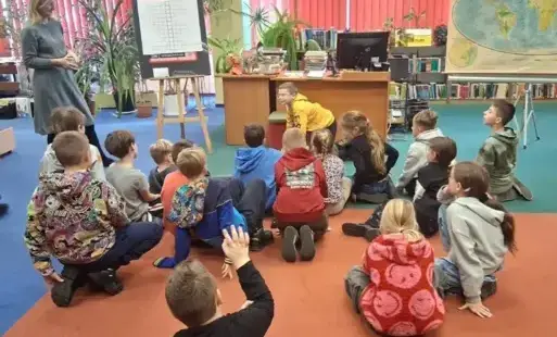 Uczniowie siedzą na wykładzinie. Przed nimi chłopiec w żółtej bluzie pokazuje wylosowane zwierzę. Po lewej stronie stoi bibliotekarka.