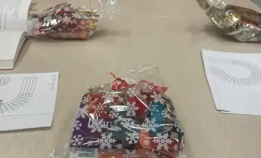 Długi stół, widoczne dłonie, kartki, długopisy, słodkie upominki świąteczne w przezroczystych małych torebkach.