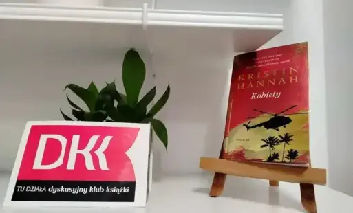 Książka „Kobiety” Kristin Hannah stoi na małej sztaludze na białej półce. Obok niej jest logo DKK i mała liściasta roślina.