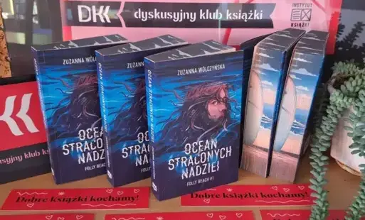 Na tle plakatu DKK stoją książki „Ocean straconych nadziei”, trzy przodem - ukazując okładkę, dwie bokiem – prezentując barwione brzegi książki.