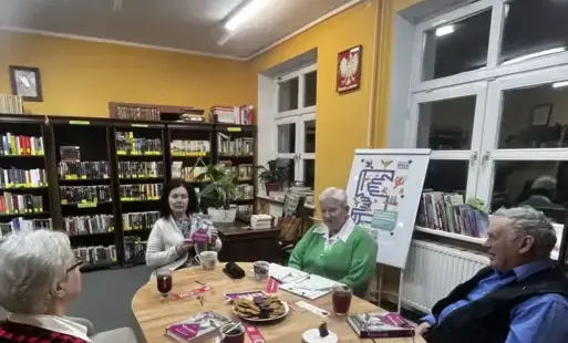 Klubowicze w rozmowie na temat książki, w tle regały na książki, oraz logo DKK.