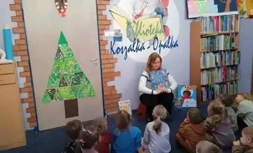 Kobieta czyta książkę grupie dzieci siedzących przed nią na dywanie