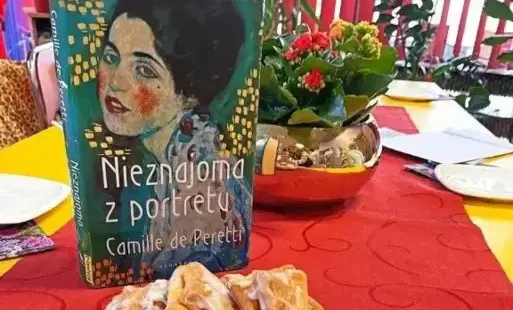 Na pierwszym planie jest widoczny talerz z różnymi słodyczami i ciastkami, zaraz obok stoi książka
