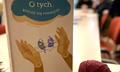 Książka stoi na długim stole, obok kłębek bordowej wełny. W tle siędzą dorośli.