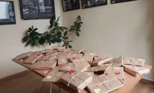 Na pierwszym planie stół  z paczkami zapakowanymi w papier koloru słomkowego, paczki obwiązane są sznurkiem na nich czerwone różowe i żółte serduszka. W tle jasno-żółta ściana na niej czarno-białe obrazy i zielona roślina.