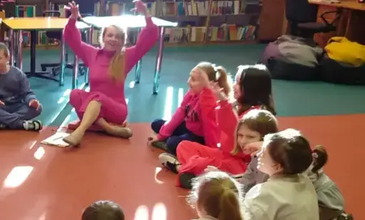 Aktorka z ekspresją i rękoma w górze opowiada, a dzieci na nią patrzą. Wszyscy siedzą na dywanie.