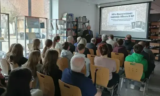 Mężczyzna stoi przy ekranie naprzeciwko siedzących ludzi.