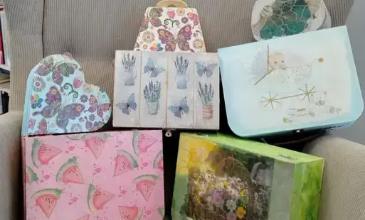 Beżowy fotel stoi na środku. Drewniane kuferki ozdobione przez uczestniczki zajęć „Pamiątki w stylu vintage” leżą na fotelu. W tle widać kawałek drzwi i białej ściany, kawałek komody.