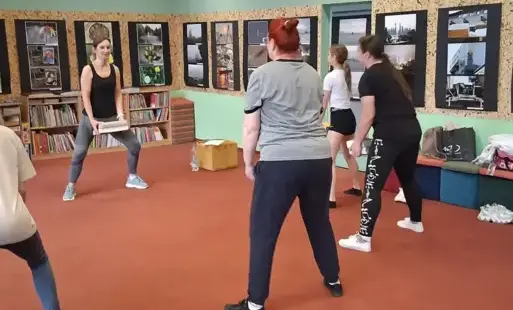 Uczestniczki zajęć stoją w rozkroku i wykonują przysiady sumo, trzymając w rękach książki jako obciążenia.
