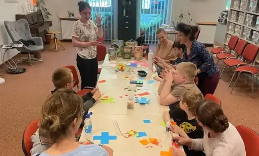 Widok na salę z długim stołem, przy którym dzieci wraz z opiekunkami i bibliotekarką wykonują pracę plastyczną.