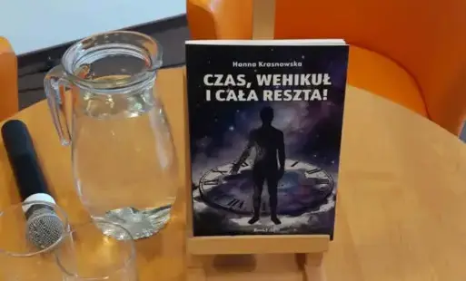 Na stojaku stoi książka 'Czas, wehikuł i cała reszta'. Po lewej stronie stoi dzbanek z wodą oraz leży mikrofon.