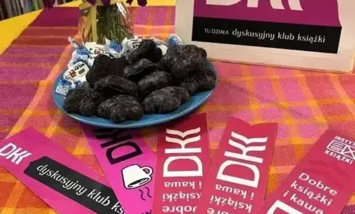 na stole ułożone są zakładki z logo Dyskusyjnego Klubu Książki, w tle ciastka i kwiatki
