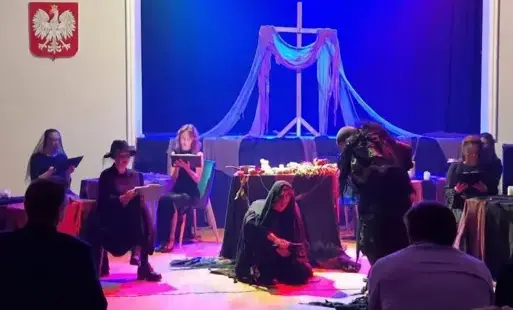Aktorzy w czarnych strojach na scenie podczas spektaklu z dekoracją w kształcie krzyża.
