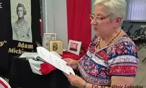 Kobieta czyta z kartek na tle polskiej flagi i portretu wieszcza.