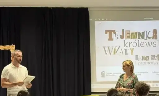 Dwie osoby przemawiają do zgromadzonej publiczności na tle ekranu z prezentacją.
