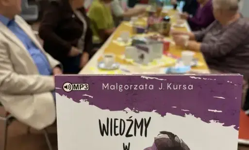 Na pierwszym planie okładka audiobooka o tytule Wiedźmy w opałach Małgorzaty J. Kursy. W tle długi stół przy którym siedzi osiem dorosłych osób.