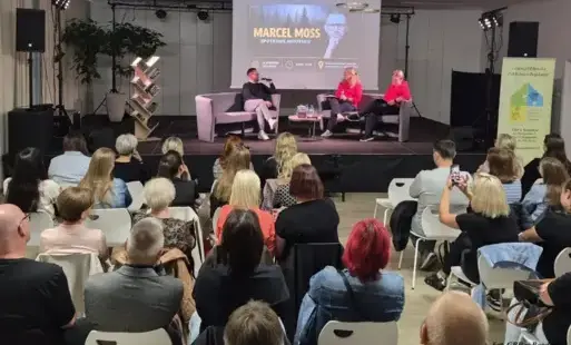 Na scenie zasiadają trzy osoby, przed scena siedzą tyłem słuchacze.