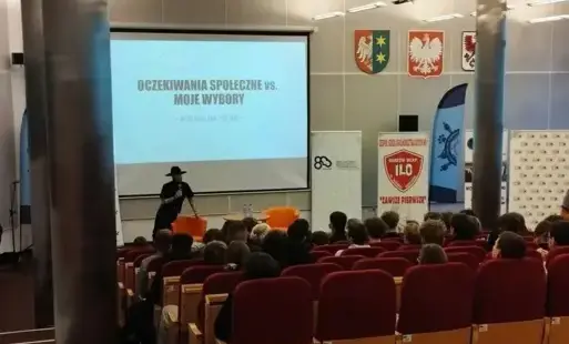 Prowadzący konferencję zwraca się do publiczności. W tle, na ekranie, widoczna jest prezentacja z tytułem „Oczekiwania społeczne vs. moje wybory”. 