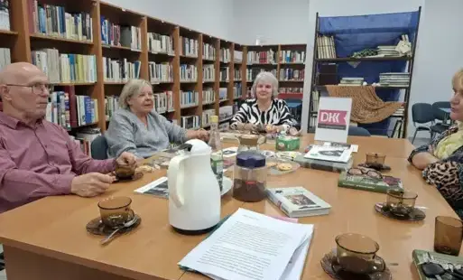 cztery dorosłe osoby siedzą na tle ściany zastawionej regałami z książkami; na pierwszym planie na stole są kartki z wydrukowanym tekstem, książki i serwis do kawy; na drugi planie na stole jest tabliczka z logo programu DKK