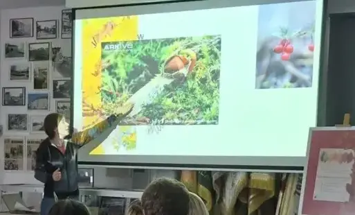 Kobieta stoi przy ekranie projekcyjnym i wskazuje dłonią na wyświetlaną prezentację multimedialną, która przedstawia zdjęcia przyrodnicze, a grupa słuchaczy siedzi przy stole konferencyjnym, tyłem lub bokiem do obiektywu, i uważnie ogląda pokaz.