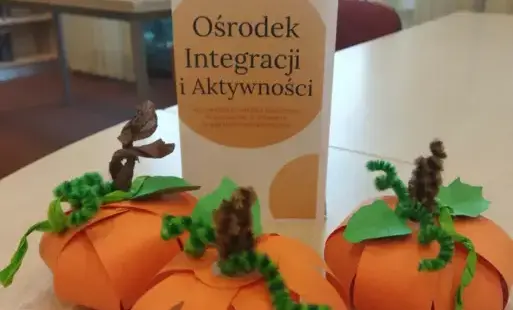 ulotka z nazwą Ośrodka, przed nią papierowe, pomarańczowe dynie