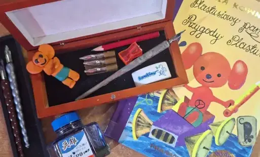 Na biurku leży książka „Plastusiowy pamiętnik”, drewniany piórnik z Plastusiem, piórem, ołówkiem, gumką i temperówką oraz buteleczka z atramentem i stalówki.