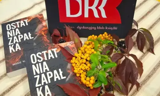 W kompozycji tytułowej omawiane książki leżą na stole po lewej stronie. Powyżej jest logo DKK. Po prawej jest jesienna kompozycja z liści i żółtych owoców irgi.
