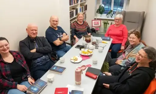 Grupa siedmiu osób siedzi przy podłużnym stole podczas spotkania Dyskusyjnego Klubu Książki w bibliotece. Na stole leżą egzemplarze książki „Śnieg przykryje' Michała Śmielaka, kubki z napojami i słodkie przekąski. W tle widać regały pełne książek oraz okn