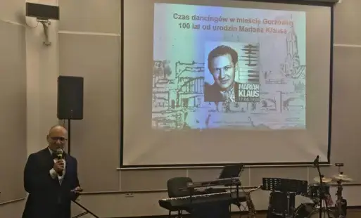 Mężczyzna przemawia przed publicznością, w tle wyświetlany slajd.