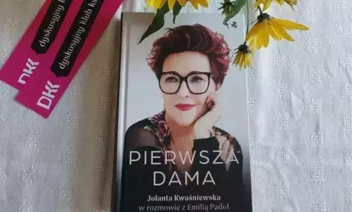Na kremowej serwecie książka, fioletowe emblematy Dyskusyjnego Klubu Książki, bukiet żółtych kwiatów.