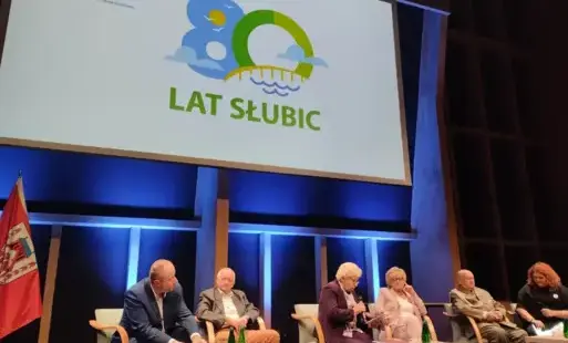 Paneliści siedzący na scenie. Za nimi wyświetla się logo obchodów 80-lecia Słubic