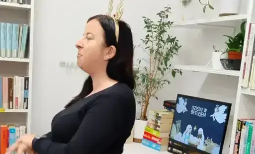 Prowadząca zajęcia, Agata Zawadzka pokazuje jak wymigać słowa kolędy. Za nią jest białe biurko, monitor z wyświetlonym slajdem kolędy, książki i białe półki, na których stoją rośliny.