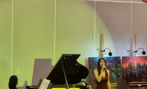 Sala audytoryjna biblioteki. Na pierwszym planie widać głowy uczestników koncertu. Na scenie na tle podświetlonej na zielony kolor ściany stoi fortepian, na którym gra pianistka. Obok fortepianu stoi wokalistka. W dłoni trzyma mikrofon. Za nią na sztaluga