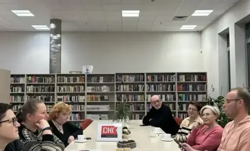 Na zdjęciu widać uczestników spotkania, którzy siedzą przy dużym, prostokątnym stole. Na stole stoją filiżanki z herbatą i kawą oraz ciastka i pierniki. Z tyłu znajdują się regały z książkami. 