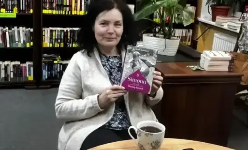 W nętrze biblioteki. Przy stole siedzi kobieta i trzyma w dłoniach omawianą książkę