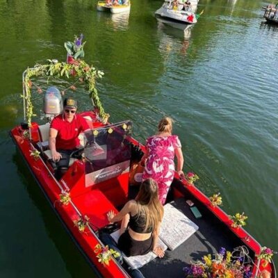 Na pierwszym planie czerwona łódź udekorowana roślinami, w tle jezioro i drzewa.