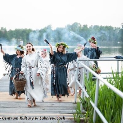Grupa tańczących osób schodząca z mostu nad jeziorem. Wszyscy przebrani w stroje na Noc Świtojańską.